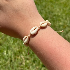 May include: A natural-toned seashell bracelet. The bracelet features several small, cream-colored seashells strung together on a beige cord. The bracelet is shown on a person's arm against a blurred green background.