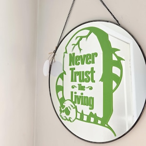 May include: A round wall mirror with a thin black metal frame and a black chain for hanging. The glass features a large green graphic of a tombstone with the text Never Trust The Living and a skull at the base, with two small white circular tags.