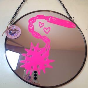 May include: Circular mirror with a black frame and chain. A neon pink graphic of a chain, hearts, and a spiky shape is on the mirror's surface. A tag with text is attached to the chain.