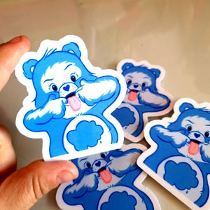 May include: A selection of blue Care Bear stickers. Each sticker depicts a blue bear with a white face, a pink tongue, and a cloud-shaped belly. The stickers are held in a hand.