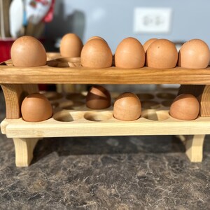 May include: A two-tiered wooden egg holder with a light brown finish. The top tier holds several brown eggs, while the bottom tier has additional egg slots. The holder is made of light-colored wood with a sturdy design.