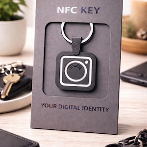 Puede incluir: Un llavero NFC en blanco y negro con un diseño cuadrado, sujeto a un llavero plateado. El llavero se muestra en una caja de cartón gris con el texto "NFC KEY" y "YOUR DIGITAL IDENTITY".