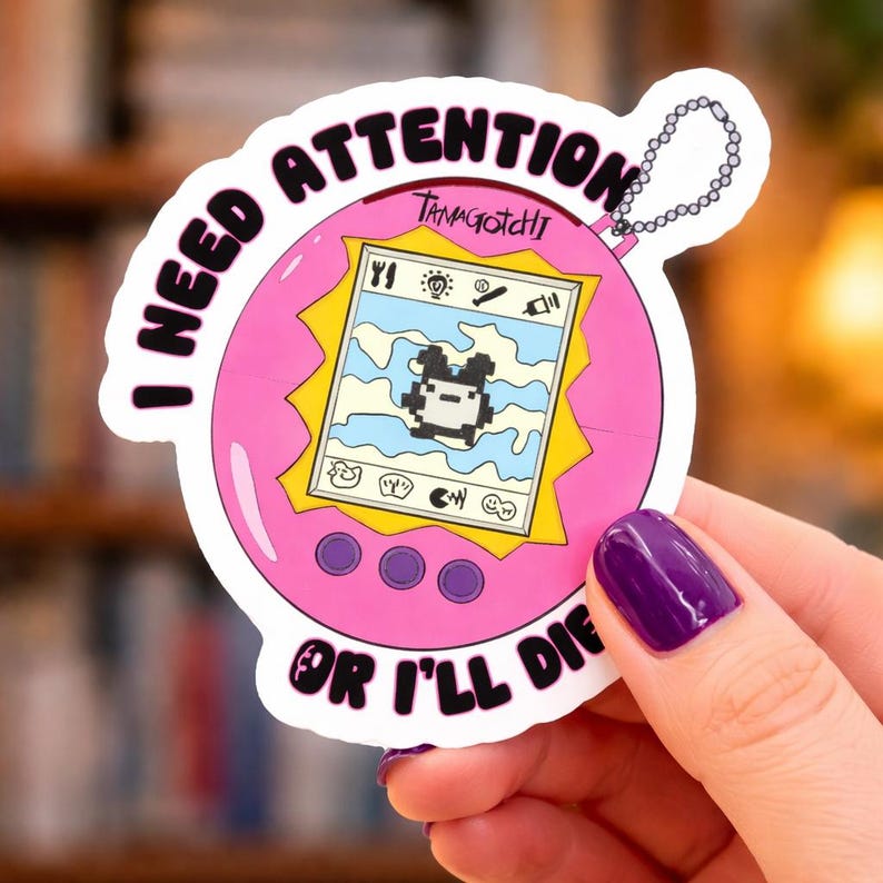 May include: A pink Tamagotchi sticker with the text "I NEED ATTENTION OR I'LL DIE." The sticker features a pixelated character on the screen. The word "Tamagotchi" is written above the screen.