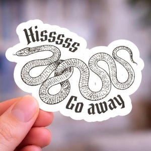 May include: A white sticker featuring a black illustration of a snake with the words "Hissssss Go away" in a gothic font. The snake is coiled and detailed. The sticker is held by a hand.