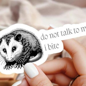 May include: A black and white sticker featuring an opossum illustration. The opossum is surrounded by the text "do not talk to me i bite". The sticker has a white background.