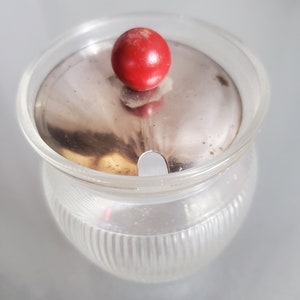 May include: A clear glass sugar bowl with a ribbed design. It has a stainless steel lid and a bright red, round knob on top. The bowl is empty and sits on a reflective surface.