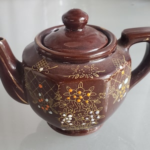 May include: A brown ceramic teapot with a lid and handle. The teapot is decorated with gold and white floral patterns. The spout and handle have gold trim. A small, round knob sits atop the lid. The teapot is likely vintage.