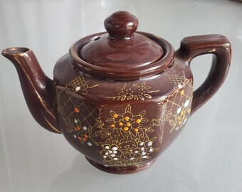 May include: A brown ceramic teapot with a lid and handle. The teapot is decorated with gold and white floral patterns. The spout and handle have gold trim. A small, round knob sits atop the lid. The teapot is likely vintage.