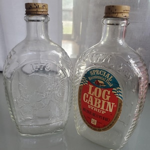 May include: Two clear glass Log Cabin Syrup bottles with cork stoppers. One bottle has an embossed design and the number "1776". The other bottle has a red, white, and blue label that reads "LOG CABIN SYRUP". Each bottle holds 24 fl oz.