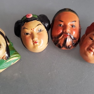 May include: Four small ceramic figurines with painted faces. The figurines are of a woman with a pink flower in her hair, a man with a beard, a woman with a red hat, and a small animal with green leaves.