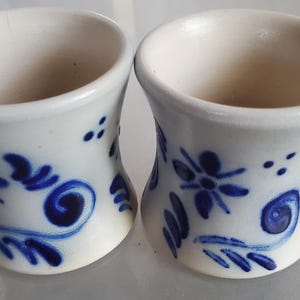 May include: Two white ceramic egg cups with blue floral designs. The cups have a flared shape and are decorated with blue flowers and swirls.