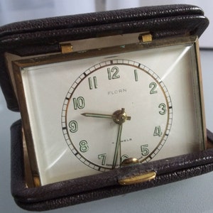 May include: A vintage travel alarm clock in a brown leather case. The clock face is white with black numerals and hands, and the brand name "FLORN" is visible. The case is open, revealing the clock face and gold-colored metal trim.