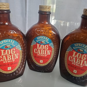 May include: Three brown glass bottles of Log Cabin Syrup. Each bottle has a label with the text "Special Bicentennial Flask" and "Log Cabin Syrup". The bottles are filled with syrup and have cork stoppers.