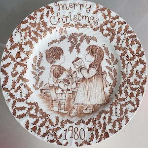May include: A decorative plate with a brown and white design. The plate features a central illustration of three children with gifts, the words "Merry Christmas", and the year "1980". The border is decorated with holly leaves and candy canes.