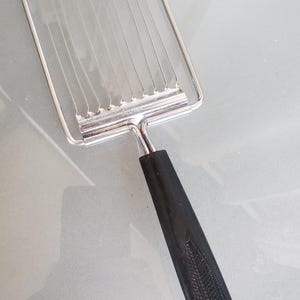May include: A silver wire-frame food slicer with a black plastic handle. The handle has a textured pattern.