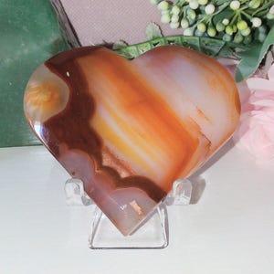 May include: A heart-shaped agate stone with bands of brown, orange, and white. The polished stone sits on a clear acrylic stand. Pink roses and greenery are in the background, suggesting a decorative or collectible item.