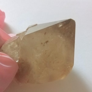 May include: A translucent, light brown crystal with a faceted, geometric shape. The crystal has a smooth, polished appearance and is held by a pink gloved hand. The crystal's color ranges from pale yellow to a deeper amber hue.