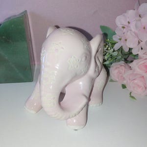 May include: A light pink ceramic elephant figurine with embossed floral patterns. The elephant has a trunk, ears, and four legs. The figurine is on a white surface. Decorative item.