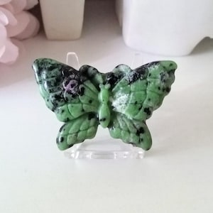 May include: A carved butterfly figurine in shades of green, black, and white. The butterfly is made of a stone-like material and is displayed on a clear stand. The wings have a speckled pattern, and the body is a lighter green.