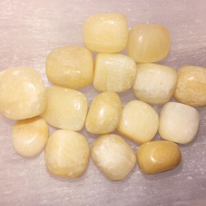 May include: A collection of smooth, tumbled yellow calcite stones. The stones vary in shades from pale yellow to a deeper golden hue. They are arranged on a light-colored, possibly marble, surface.