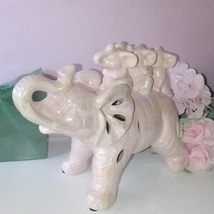 May include: A ceramic figurine of a light pink elephant with three smaller elephants on its back. The elephant has cut-out details on its ears and legs. The figurine is on a white surface with pink flowers in the background.
