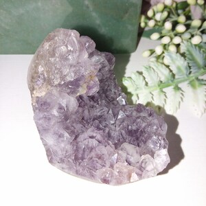 May include: A cluster of amethyst crystals in shades of purple and lavender. The crystal formation has a rough, textured surface with many small, pointed crystal structures. The amethyst is displayed on a white surface with green and white decorative elements in the background.