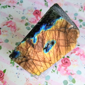 May include: A triangular-shaped labradorite gemstone with a mix of colors. The stone features a dark gray top, transitioning to blue and yellow hues. The surface has visible cracks and a polished appearance, showcasing its natural beauty.
