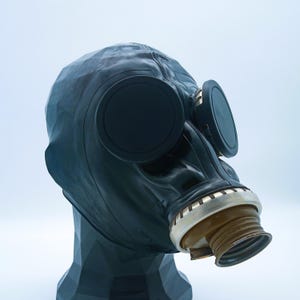 Könnte beinhalten: Eine schwarze Gasmaske mit großen, kreisförmigen Okularen und einem Gewinde-Mundstück. Die Maske besteht aus einem dunklen, gummiartigen Material und ist auf einem schwarzen, geometrischen Ausstellungsständer montiert. Die Okulare sind schwarz und das Mundstück ist beige.