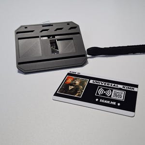 Könnte beinhalten: Ein schwarzer Ausweishalter mit einem schwarzen Schlüsselband und einer weißen Karte. Die Karte trägt den Text "UNIVERSAL KINK" und "SCAN ME" mit einem QR-Code. Der Ausweishalter hat einen Schiebemechanismus.