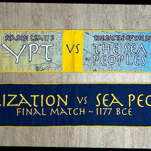 May include: Two blue and yellow fabric scarves with fringe. The top scarf has the text "EGYPT VS THE SEA PEOPLES" and the bottom scarf has the text "CIVILIZATION VS SEA PEOPLES FINAL MATCH ~ 1177 BCE".
