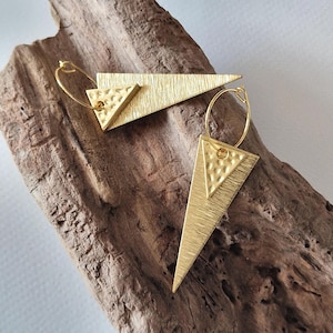 May include: Gold-coloured earrings with a geometric design. Each earring has a large, textured triangle with a smaller triangle overlay and a hoop closure. The earrings are displayed on a piece of driftwood.