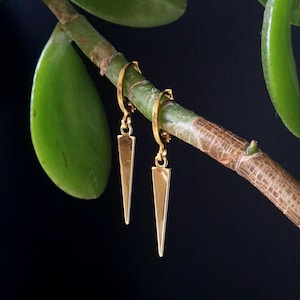 May include: A pair of gold-coloured earrings with a triangular pendant design. The earrings are suspended from a green plant stem against a black backdrop. The earrings are small and delicate, with a polished finish.