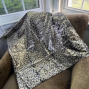 May include: A square fabric scarf featuring a repeating leopard print pattern in shades of black, grey, and metallic gold. The material has a reflective, shimmering finish that catches the light, creating a high-contrast animal print design.