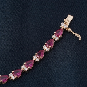 May include: A gold bracelet with alternating pear-cut, deep red gemstones and clusters of small, clear stones. The bracelet is secured with a gold clasp, displaying an elegant design.