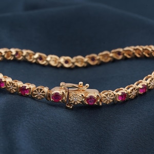 Op de afbeelding: Goudkleurige armband met rode edelstenen in bloemvormige schakels. De armband heeft een veilige sluiting met een kleine diamant. De sieraden worden gepresenteerd tegen een donkerblauwe achtergrond.