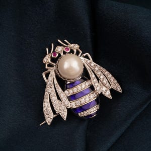 Puede incluir: Broche de abeja plateada con cuerpo de perla grande, abdomen a rayas azules y blancas y detalles de diamantes en las alas. Los ojos de la abeja son pequeñas gemas rojas. Sobre tela azul oscuro.