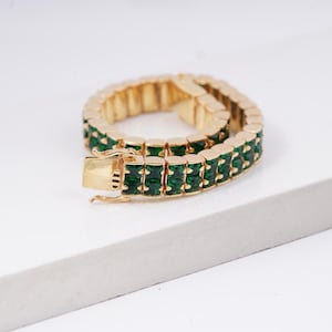 Könnte beinhalten: Goldfarbenes Armband mit rechteckigen, smaragdgrünen Edelsteinen. Das Armband hat ein zweireihiges Design mit einem sicheren Verschluss. Die Edelsteine sind in einen goldfarbenen Metallrahmen gefasst, was einen luxuriösen Look erzeugt. Das Armband wird auf einer weißen Oberfläche präsentiert.