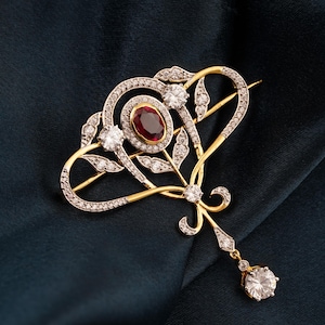 Könnte beinhalten: Gold- und Silberbrosche mit herzförmigem Design. Die Brosche hat einen zentralen ovalen Rubin, umgeben von Diamanten und einem hängenden Diamantanhänger. Die Brosche ist vor einem dunkelblauen Hintergrund platziert.