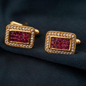 以下が含まれることがあります： 長方形のデザインの金色のカフスボタンのペア。各カフスボタンは、小さな正方形の濃いピンク色の宝石の集まりを特徴とし、小さな透明な宝石の境界線で囲まれています。外側のフレームは金色です。