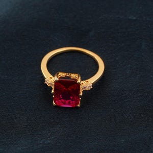 May include: A gold-colored ring featuring a large, rectangular, deep red gemstone as the centerpiece. The ring has small, clear accent stones on either side of the main stone, adding sparkle. The band is a simple, polished gold.