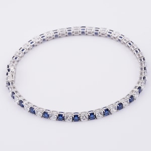 May include: A silver bracelet featuring alternating blue and clear square-cut gemstones. The bracelet is designed with a classic tennis bracelet style, showcasing a continuous line of sparkling stones. The clasp is visible, indicating a secure closure.