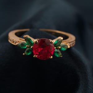 Peut inclure: Une bague dorée ornée d'une grosse pierre rouge ronde au centre. La bague est ornée de petites pierres vertes et de minuscules pierres transparentes le long de la bande, sur fond bleu foncé.