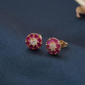 May include: A pair of gold stud earrings. Each earring has a flower-like design, with a ring of small, red gemstones around a central cluster of smaller, clear gemstones. The earrings are displayed on a dark blue background.