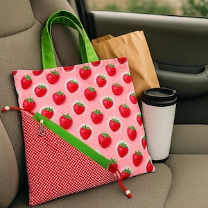 May include: A pink tote bag featuring a strawberry design, green handles, and a red polka dot pocket. A white travel mug with a black lid and a brown paper bag are also present.