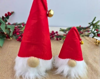 Può includere: Due decorazioni natalizie a forma di gnomo. Ogni gnomo ha un cappello conico rosso con una campana dorata, una barba bianca e soffice e un naso beige. Gli gnomi sono su una superficie beige con bacche rosse e foglie verdi sullo sfondo.