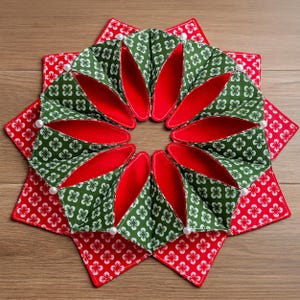 May include: A handmade, decorative star-shaped item. The design features a central ring of green fabric with white floral patterns and red interiors, surrounded by red fabric squares with white floral patterns. White pearl-like beads are placed around the edges.
