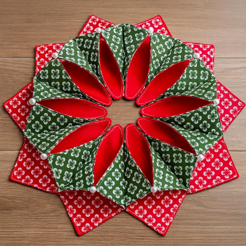 May include: A handmade, decorative star-shaped item. The design features a central ring of green fabric with white floral patterns and red interiors, surrounded by red fabric squares with white floral patterns. White pearl-like beads are placed around the edges.