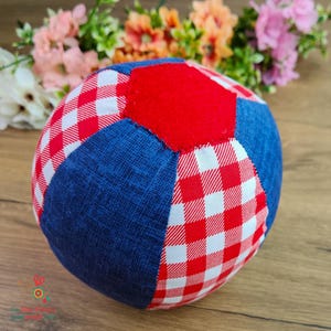 May include: A round, fabric ball with a patchwork design. The ball features red and white gingham check patterns, solid red panels, and blue denim-like fabric sections. The ball is designed for play.