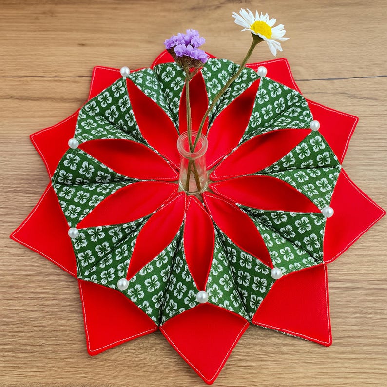 May include: A decorative, star-shaped table centerpiece with a red base and green and white patterned petals. The petals are adorned with white pearls. A small glass vase holds purple and white flowers, adding a touch of natural beauty to the handcrafted design.