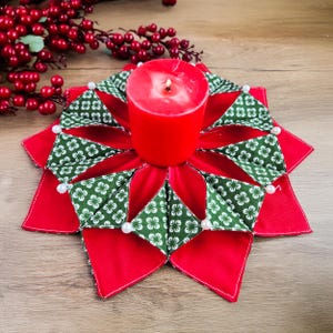 May include: A red pillar candle sits atop a handmade candle holder. The holder is a starburst design with red fabric and green fabric with a white floral pattern. White pearls are sewn on the points of the star.